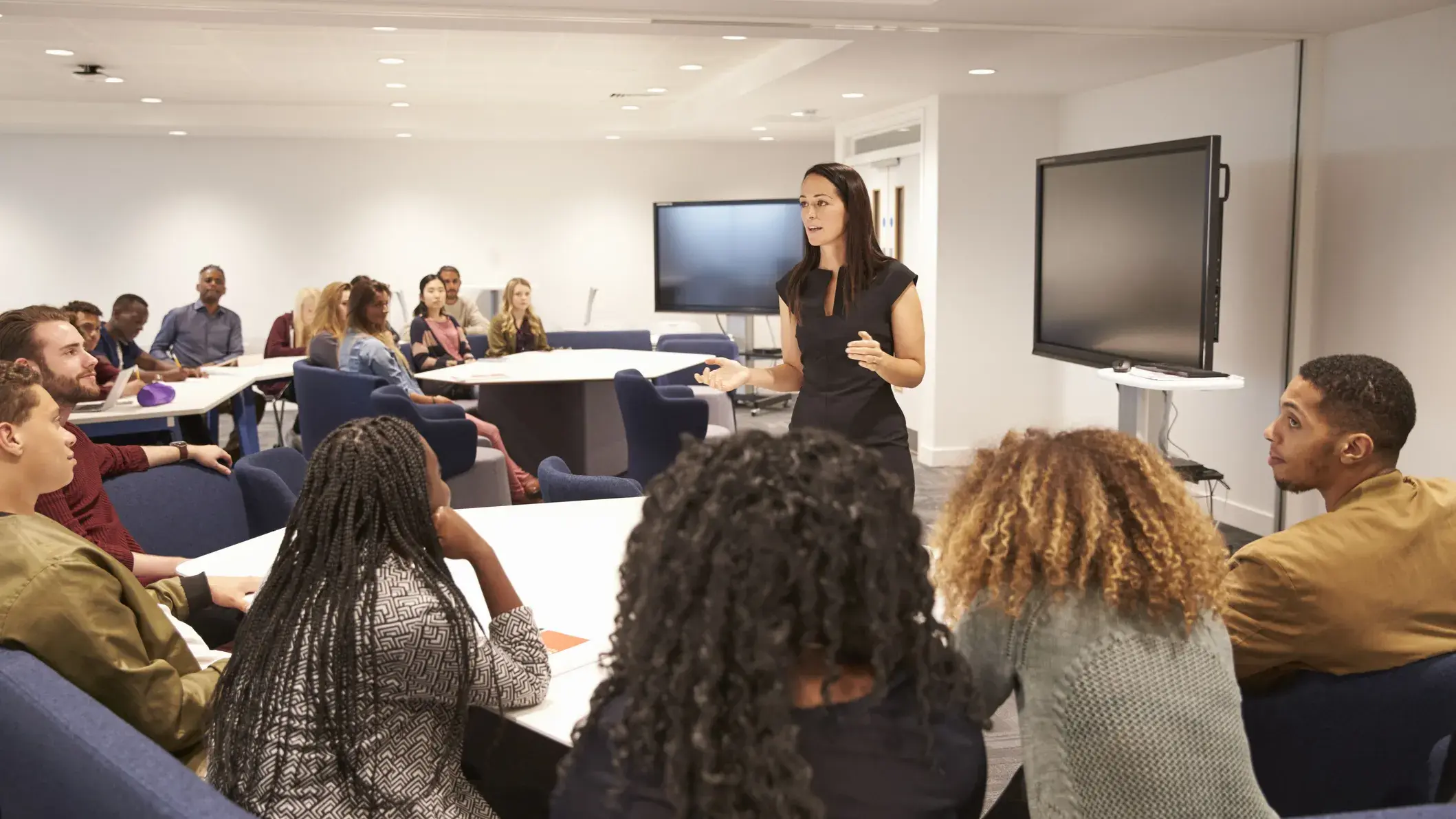 woman in a black dress speaking in front of a group of attentive people in a classroom setting