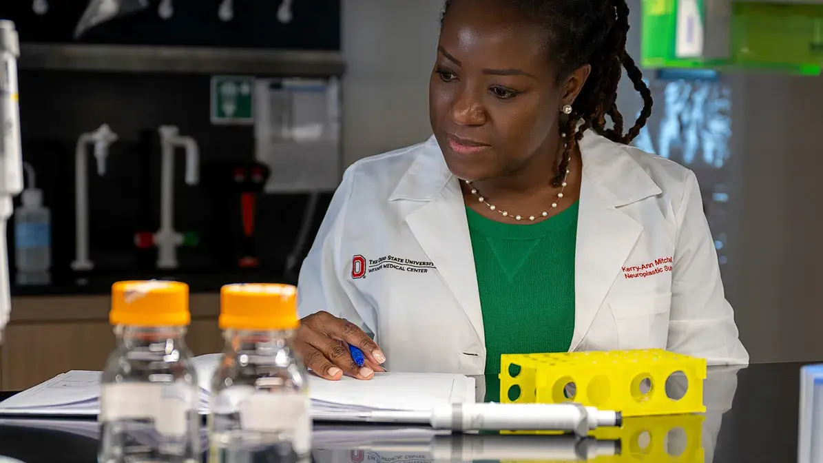 woman in Ohio State lab coat sitting in a lab writing in a notebook