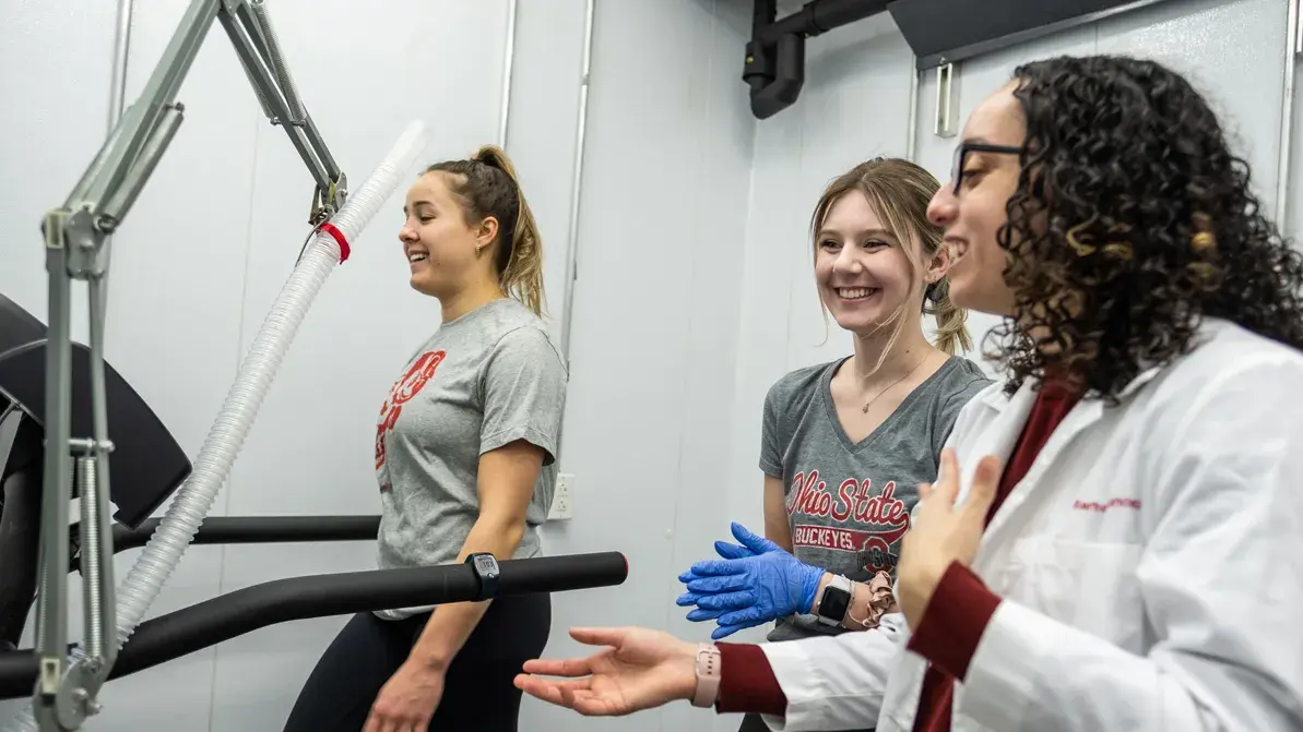 woman in lab coat explaining a testing treadmill machine to two women